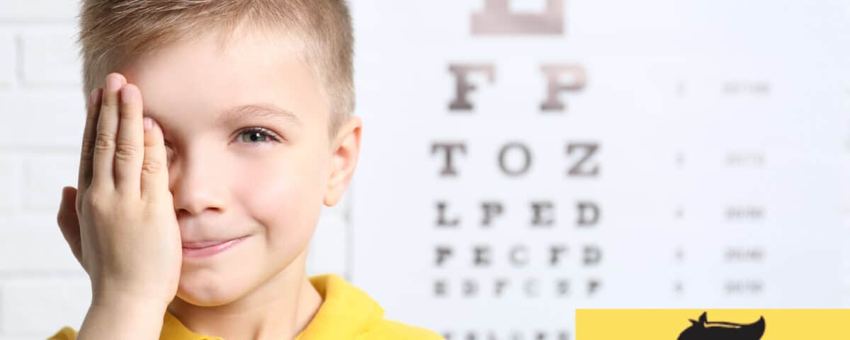 kid holding eye with hand with a blurred out eye exam behind him.