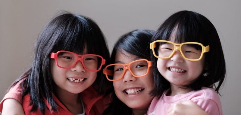 Three smiling children with straight black hair are wearing large, colorful glasses—red, orange, and yellow—while posing closely together in front of a plain background.