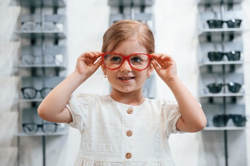 A young child wearing a white dress smiles while trying on oversized red eyeglasses in an eyewear store, with display shelves of glasses blurred in the background.